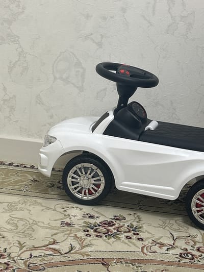 A white toy car with a black steering wheel sits on a patterned rug. The car has silver rims on its wheels and red accents visible through the spokes. The background is a light-colored wall with a textured pattern. The image does not provide any information about the city of Buynaksk, Russia.