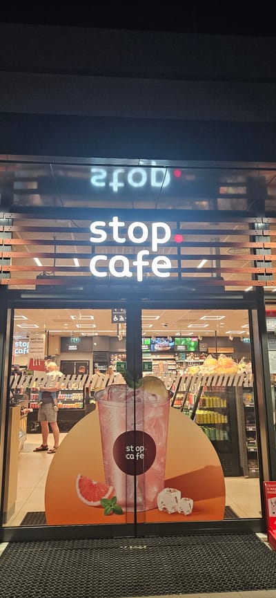 This image shows the entrance to a Stop Cafe convenience store located in Sobieszczany-Kolonia, Poland. The store's sign, illuminated with white lettering, reads "stop cafe" with a red dot serving as the 'o' in "stop" and another red dot to the right of "cafe." Above the main sign, a reversed reflection of the store's name is visible.

The entrance consists of glass double doors, through which the interior of the store is visible. Inside, shelves are stocked with various products, and a counter area is present. A man is standing near the entrance, holding a child, and other customers are visible inside the store. A large advertisement featuring a drink with ice, mint, and grapefruit slices is displayed on the glass doors. Additionally, accessibility symbols (wheelchair and person with cane) are posted near the entrance. The overall environment appears to be nighttime, indicated by the artificial lighting.