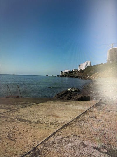 The image depicts a waterfront scene with a concrete walkway in the foreground. To the left of the walkway, a set of metal steps leads into the calm, blue water. The water extends to the horizon, meeting a clear blue sky. Along the coastline in the distance, several white buildings are visible on a hillside, with a construction crane towering over one of them. The environment suggests a sunny day at a coastal location. The image does not provide any information about the city of Mirnoye, Ukraine, or any specific details about people or actions.
