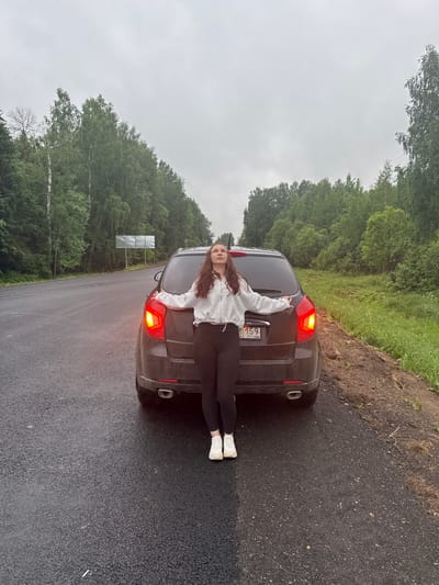 A long-haired woman in a gray hoodie and black leggings stands behind a dark-colored car parked on a wet asphalt road in a wooded area. The car's taillights are illuminated. The car's license plate is visible, displaying "159" and the letters "RUS" indicating the country of registration. In the background, dense green trees line the roadside under an overcast sky. A signpost is partially visible in the distance. The location is in the city of Михайловка, Russia.