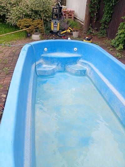 The image depicts an empty, bright blue plastic wading pool filled with a shallow layer of murky water. The pool is situated outdoors on a paved surface made of reddish-brown bricks. In the background, there is a collection of items including a yellow and grey pressure washer with the number "28" visible, a small yellow toy scooter, and a black and yellow cleaning machine. Beyond these objects, lush green foliage and trees are visible, with a wooden fence in the far background. The scene appears to be a backyard or garden area, possibly in Zawiercie, Poland, given the context. There are no people visible in the image.