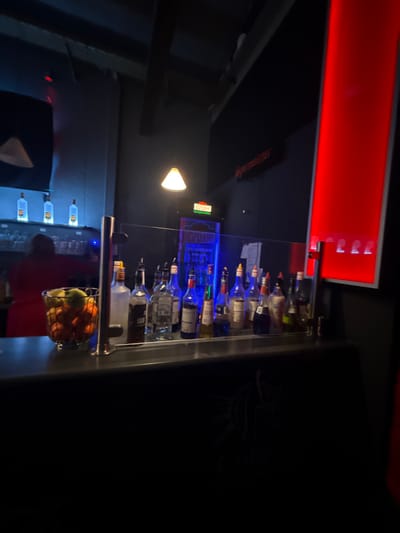 A bar with multiple alcohol bottles lined up behind a transparent partition with metal railings. Above, there is a red illuminated panel. Below, vodka bottles glow blue. A hanging basket holds oranges and apples. A lamp casts light onto the bar area. A dark figure in a red shirt stands near the bar, seemingly engaging with it.
