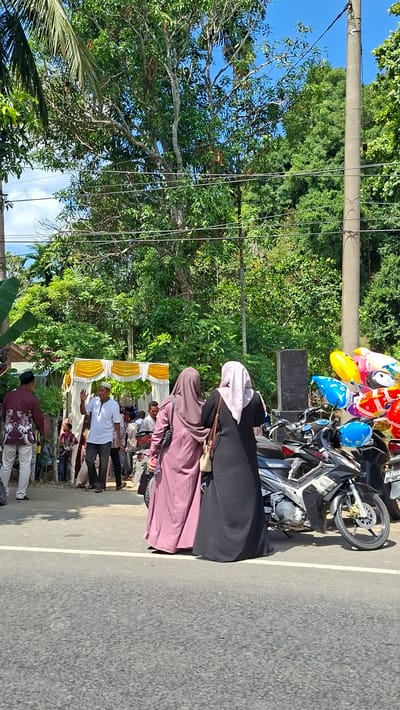 Two women in hijabs, one in a dusty rose colored abaya and the other in a black abaya, are standing on the side of a road in Sawang, Indonesia. They are facing away from the camera, with the woman in the black abaya holding a light beige purse on her right shoulder. To their right, parked on the shoulder, is a black motorcycle. Behind the motorcycles, a cluster of colorful balloons are tied to a post. In the background, a white and yellow tent structure is set up, with several people gathered around it, some of whom are looking towards the street. Trees and foliage line the background, with a utility pole and power lines also visible. The road is paved with asphalt and has a white line marking the edge.