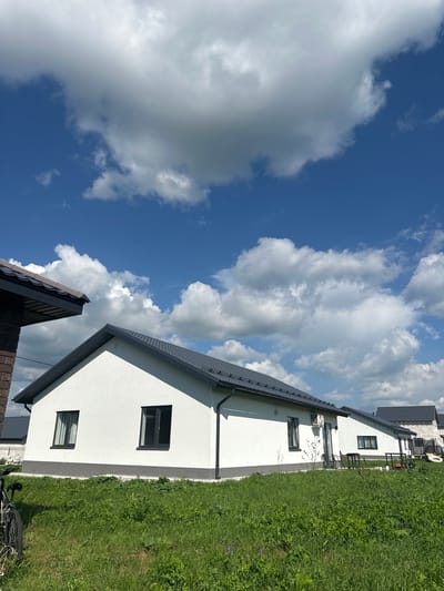 The image depicts a bright, sunny day in Mokino, Russia, with a clear blue sky dotted with fluffy white clouds. In the foreground, a lush green lawn stretches out, with a bicycle parked on the left side. Several modern, white houses with dark gray roofs are visible, some under construction. The houses are single-story structures with clean lines and dark-framed windows. One house has an air conditioning unit mounted on its exterior wall. In the background, more houses and some building materials, including what appears to be a trampoline, can be seen. The overall impression is one of a developing residential area on a beautiful summer day.