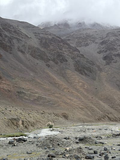 The image depicts a mountainous landscape in Sumdo, India. A wide, rocky riverbed dominates the foreground, with a shallow, fast-flowing river cutting through it. A solitary tree with light-colored foliage stands in the middle of the river, its roots embedded in a rocky island. The riverbanks are lined with grey and brown rocks of varying sizes, interspersed with patches of sparse green vegetation.

In the midground, the steep, barren slopes of a mountain rise sharply. The mountainside is textured with scree, small rocks, and evidence of past erosion, creating a rugged, uneven surface. Faint, thin lines, likely power lines, stretch horizontally across the lower section of the mountain slope, indicating human presence.

The background features more towering mountains, their peaks shrouded in thick, white clouds. The atmosphere appears overcast, casting a muted light over the entire scene and contributing to a sense of isolation and rugged beauty. No people or animals are visible in the image.