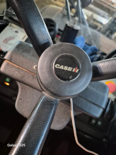 A close-up shot shows the steering wheel of a vehicle, likely a tractor, with the "CASE IH" logo prominently displayed on the center hub. The steering wheel is covered in black, textured material, possibly leather. The background is blurred, but appears to be the interior of a vehicle, with some equipment and a blue cloth visible. There are also some lights and gauges in the background. The image is likely taken in Pątnów, Poland, as indicated by the watermark "Galaxy S25" at the bottom left corner, a common characteristic of Samsung phones sold in certain regions. The scene is static, with no people or actions depicted. The overall impression is of a functional, well-used piece of agricultural or industrial equipment.