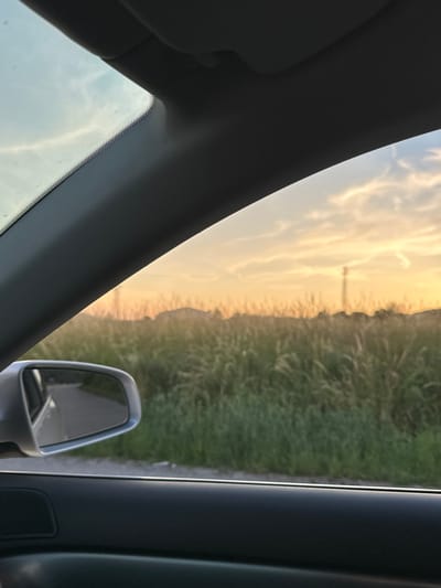 This is a photograph taken from inside a vehicle, looking out through the front windshield and side window. The perspective suggests the vehicle is moving. The upper portion of the image is dominated by the car's interior trim, likely the A-pillar and headliner, which are dark gray and textured. Through the windshield, the sky displays a soft, hazy sunset with pastel colors of orange, yellow, and pale blue, interspersed with wispy clouds.
To the right of the car's interior, the view opens up to a landscape. A vast field of tall, green grass occupies the middle ground, swaying gently. Beyond the field, a low, dark silhouette of hills or mountains can be seen against the sunset sky. A solitary utility pole is visible in the distance, standing tall against the horizon.
In the lower left quadrant of the image, a side-view mirror is prominently featured. The mirror reflects a gray asphalt road and the green grass bordering it, indicating the car is on or next to a road. The bottom of the image shows the interior of the car door, a dark, textured surface with a speaker grille visible on the left. The overall impression is one of a serene, late afternoon or early evening drive through a rural area. The location is identified as Debelets, Bulgaria.