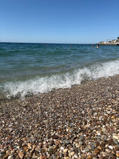 The image shows a pebble beach with the sea in the background under a clear blue sky. A gentle wave is breaking on the shore. In the distance, several people are swimming in the water. To the right, a white building with an arched structure is visible on a rise overlooking the sea. The scene is likely located on a sunny day in Кореизский поселковый совет, Ukraine, given the presence of a beach and sea.