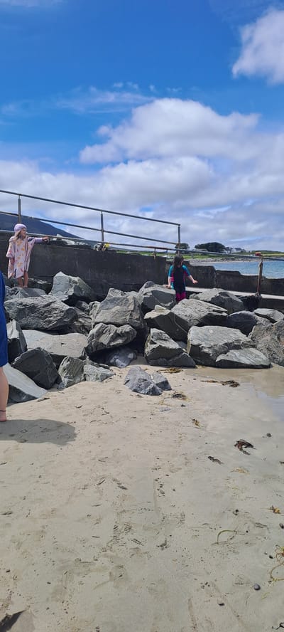 The image shows a view of a rocky pier and a sandy beach on a sunny day. There are three young people visible. The person in the foreground is partially obscured, wearing a pink patterned top and a light-colored head covering. Another person, dressed in a black and pink swimsuit, is walking on the rocks. The third person is not clearly visible. The sky is blue with fluffy white clouds, suggesting fair weather. The presence of a pier, the ocean in the background, and the rocky and sandy terrain indicate a coastal setting, consistent with the provided location context of Pier Road, Ireland. The scene appears to be a casual day out at the beach, with children exploring the environment. The sun is casting shadows, indicating it is likely daytime. There is no visible text in the image.