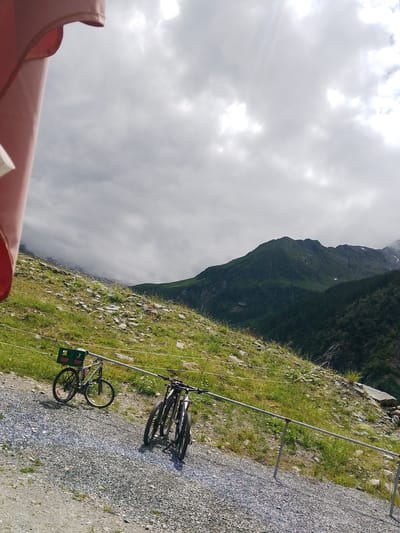 The image shows a scenic mountain landscape in Bramberg am Wildkogel, Austria. In the foreground, two bicycles are parked on a gravel path. One bicycle, positioned further left, is a silver mountain bike with a green beer crate strapped to its rear rack. The crate displays the text "Egger" and "Gleisdorf" in white lettering. The second bicycle, to the right, is a dark-colored mountain bike with its handlebars angled. Both bicycles appear to be parked or waiting.
The foreground path leads up a grassy, rocky slope dotted with small wildflowers. The slope is bordered by a metal railing. In the background, lush green mountains rise dramatically, with darker, forested slopes in the mid-ground. The sky is overcast with dramatic, cloudy formations, suggesting a day with mixed weather. Sunlight filters through the clouds, illuminating parts of the landscape. The overall atmosphere is serene and outdoorsy, evoking a sense of adventure and natural beauty. The time of day appears to be daytime, likely late morning or afternoon, given the lighting conditions. There are no people visible in the image, implying a moment of quiet contemplation or a pause in outdoor activity.