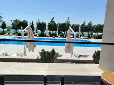 The image depicts an outdoor scene at a resort in Konigil, Uzbekistan, on a clear, sunny day. In the foreground, a dark railing obscures the lower part of the view. Beyond the railing, a tiled patio area leads to a large, bright blue swimming pool. Several closed, light-colored umbrellas are positioned around the pool. Some lounge chairs are visible on the patio, and a small table with a rounded wooden top is partially visible on the far right.

The middle ground features a row of lush green trees separating the pool area from a body of water, likely a lake or sea, in the background. On the far side of the water, a cluster of buildings with blue rooftops can be seen. The sky is a clear, pale blue with faint power lines stretching across the upper portion of the image. A few individuals are present in the scene; one person is standing near the edge of the pool in the middle right, and several others are visible near the water in the distance. The overall impression is of a tranquil resort environment on a warm day.