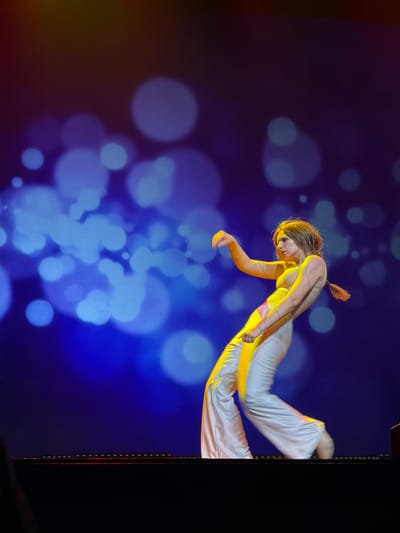 An eye-level, medium shot captures a female aerialist performing against a backdrop of glowing blue and purple bokeh lights in Ventspils, Latvia. The scene is dark, suggesting a nighttime performance or a darkened stage. The aerialist, positioned in the center, is dressed in a light-colored, possibly white or cream, leotard or bodysuit and flowing wide-leg pants. Her outfit is illuminated by a warm, golden-yellow light source from the right, highlighting her left side and casting her right side in shadow. She has light-colored hair, tied in a high ponytail that is visible behind her head. A pattern of thin blue lines is painted on her arms and possibly other parts of her body, and some glitter or reflective makeup is visible on her face, particularly around her eyes.

The aerialist is in mid-air, horizontally suspended, with her body angled slightly downward. Her left arm is extended forward and slightly bent at the elbow, hand open. Her right arm is also bent at the elbow, hand seemingly grasping something or posed. Her legs are extended, and her wide-leg pants billow around them, suggesting movement.

To the right edge of the frame, a dark, vertical structure, possibly part of a stage rig or support beam, is visible. The glowing bokeh lights behind her create a dreamy, ethereal atmosphere, with varying sizes and intensities of circular light scattered across the blue and purple background. There is no visible text in the image.