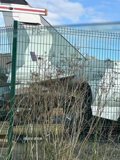 An image captured at the Manston History Museum in Thanet, United Kingdom, displays the tail section and rear fuselage of a British Aerospace Harrier GR.3 aircraft, serial number XZ106. The aircraft is painted in a light grey/white camouflage scheme with darker grey patches. Prominently visible on the tail fin are the letters "FR" and a Royal Air Force roundel on the fuselage side. The scene is viewed through a green wire mesh security fence, featuring vertical posts and horizontal stiffening ribs. In the immediate foreground, numerous dormant, leafless woody shrubs partially obscure the fence and the lower portion of the aircraft. Behind the fence, a paved ground surface is visible, upon which a large, cylindrical object, likely an external fuel tank or ordnance, rests beneath the aircraft's fuselage. The upper background consists of a clear blue sky with scattered white clouds. No people, active movements, or interactions are visible.