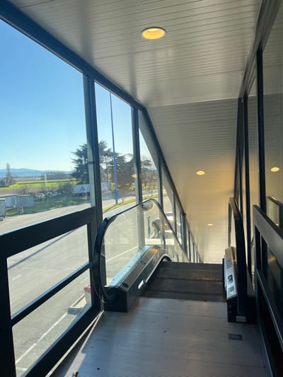 The image displays the interior of a modern building, dominated by an escalator and large windows, captured under bright, sunny conditions. The scene is oriented vertically but would show an escalator descending towards the foreground on the right side of the frame if rotated correctly. The escalator features dark steps and handrails, and a white panel with blue characters, possibly "0000," is visible near its upper section.
To the left, expansive, dark-framed windows offer a clear view of the outside world. Through the glass, a vibrant daytime landscape is visible, including green trees, a road, a parking area, and distant rolling hills or mountains under a bright blue sky. Reflections of the sky and internal elements, including what appears to be a person's silhouette, are present on the windowpanes.
The building's interior features a light-colored, paneled ceiling with multiple circular, recessed light fixtures emitting a warm light. The walls are similarly paneled. The flooring around the escalator is a dark grey. No people are actively using the escalator, and the scene is static, suggesting a quiet moment. The overall impression is of a spacious and well-lit environment, blending modern architecture with an open view of nature. The context places the location in Dorno, Italy.