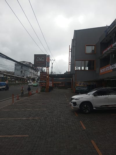 A street view features the Sturgis Tropical Hotel sign. A gray building stands adjacent, displaying "Roti Sturgis" and "Bengkel More" signs. A white SUV is parked in the lot paved with small square stones and striped with yellow lines marking parking spaces. Two people stand along the side of the road near orange traffic cones. Overhead, utility lines stretch across a cloudy sky. Other buildings are visible in the distance.