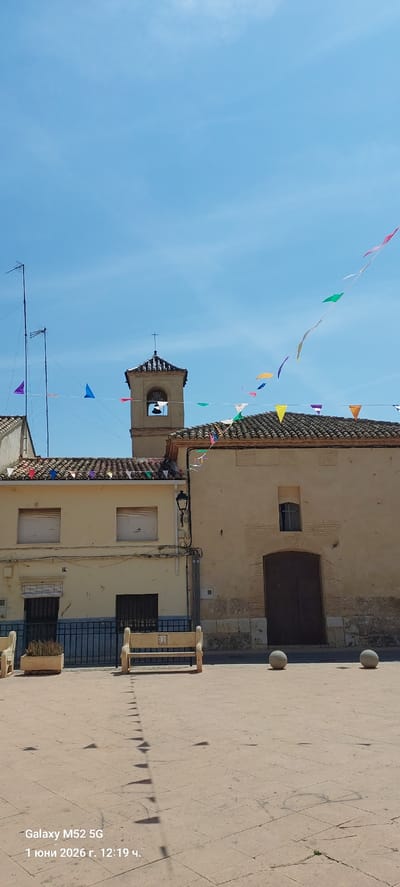 The image shows a street scene in Canals, Spain, under a clear blue sky. Buildings with beige facades and tiled roofs are decorated with colorful pennant banners strung across the street. A church tower with a cross on top is visible. The street is paved with light-colored stones. Shadows of the banners are cast on the ground. A bench and potted plants are situated along the building's edge. An antenna is mounted on a nearby roof.