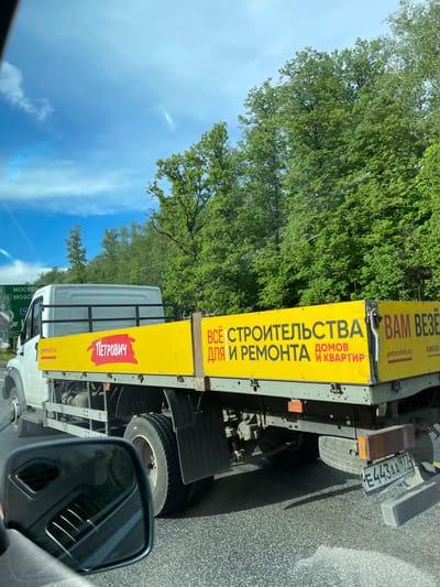 The image shows a view from inside a vehicle, focusing on a truck alongside on the road. The truck has a yellow advertising banner for "Petrovich," a construction and renovation supply company. The banner reads "Всё для строительства и ремонта домов и квартир" (Everything for construction and renovation of houses and apartments) and "Вам Везёт" (You're Lucky). The company's website, petrovich.ru, and phone number, 8 800 333-, are also displayed. The truck's license plate number is E443AA 977.
In the background, there are trees and a blue sky with some clouds. A road sign indicates "Москва 2 (Moscow 2)" and "105 MOSCO," suggesting the location is near Moscow.
Based on the license plate's regional code "977", the vehicle is registered in Moscow. The road sign indicating "Moscow 2" indicates a proximity to the city. The proximity to Moscow in the image aligns with the description of the city "Serp I Molot" as the city is also in Russia.