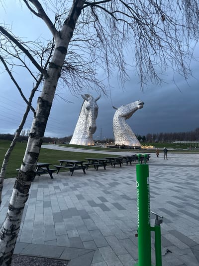 The image depicts an outdoor scene at dusk or early evening, centered on two large, illuminated equine head sculptures known as The Kelpies, located in Helix Park, Falkirk, United Kingdom. The sky is heavily overcast with dark grey clouds.
In the foreground, on the left, a silver birch tree with stark, bare branches extends diagonally from the lower left towards the top right, partially obscuring the left sculpture. Its trunk exhibits characteristic white and dark grey patterned bark. On the right side of the foreground, a bright green, cylindrical bicycle repair station stands upright, featuring "GREASEMONKEY" and "INFRASTRUCTURE" text printed vertically, along with a pump attached.
The midground consists of a paved area made of rectangular light and dark grey blocks, stretching from the bottom of the frame towards the background. On this paved surface, a linear arrangement of at least seven dark wooden picnic tables with attached benches is visible, extending towards the sculptures. To the left of the paved area, a neatly mown green lawn covers the ground.
The two prominent Kelpies sculptures, constructed from interwoven metallic plates, are situated in the background. The left sculpture presents a horse's head looking mostly forward, while the right sculpture depicts a horse's head in a rearing pose, looking upwards and to the right. Both sculptures are illuminated from their bases by warm artificial lights. Several indistinct human figures are visible walking on a path behind the picnic tables and near the bases of the sculptures. In the far background, power lines and a large pylon are discernible against the horizon, with a line of bare deciduous trees below.
