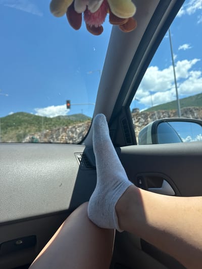 A close-up, low-angle shot focuses on a person's bare leg and foot wearing a white ankle sock, propped up inside a car. The leg is positioned against the car's interior door panel, with sunlight casting a shadow across it. The foot is bent, with the toes pointing upwards. In the background, viewed through the car window, is a bright blue sky with fluffy white clouds, lush green mountainside, and a rocky, exposed cliff face. A red traffic light is visible in the distance. The car's interior is grey and textured. The scene appears to be set in Kato Nevrokopi, Greece, possibly during daytime, as suggested by the strong sunlight and clear skies.