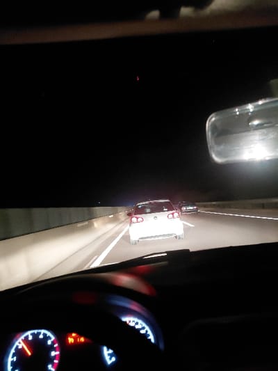 The image captures a nighttime scene from the interior of a vehicle, looking forward and slightly to the left. On the right side, parts of a steering wheel and a car's dashboard are visible. The dashboard features illuminated gauges with white markings and red needles, indicating speed, engine RPM, fuel level, and temperature. Several orange warning lights are also lit, visible near the top of the gauge cluster.

Ahead and to the left, a white car, possibly a sedan or hatchback, is traveling on an asphalt road. Its red taillights are clearly visible, contrasting with the dark surroundings. White lane markings delineate the road. To the upper left of the road, a clear glass bottle or container is illuminated, reflecting light. Further into the dark distance on the left, a small red light is faintly visible. The overall setting indicates active driving during the night.

No discernible text is visible on the dashboard or elsewhere in the scene. The image contextually places this scene in Majmaa Tolba, Morocco.