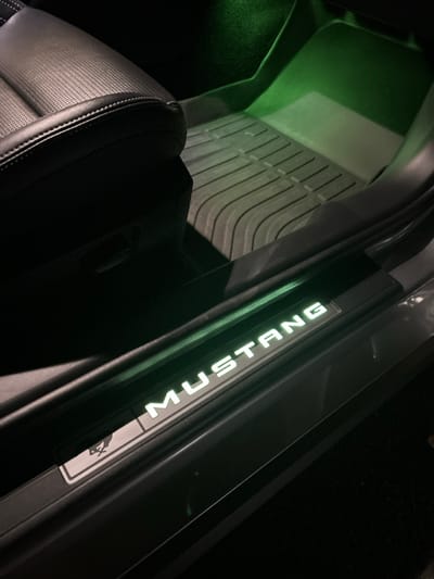 The image displays the interior passenger-side door sill and footwell of a Ford Mustang, located in Thousand Oaks, United States. The scene is illuminated by vehicle interior lights under dark conditions. A black leather car seat, featuring a perforated texture and light-colored stitching, occupies the left side of the frame. The footwell contains a dark gray, ribbed rubber floor mat. The focal point is the black plastic door sill, which features illuminated white lettering spelling "MUSTANG" and a white, illuminated Mustang pony logo on its left side. A distinct green ambient light emanates from the upper footwell area, casting a soft glow on the floor mat and lower dashboard. No individuals are visible.