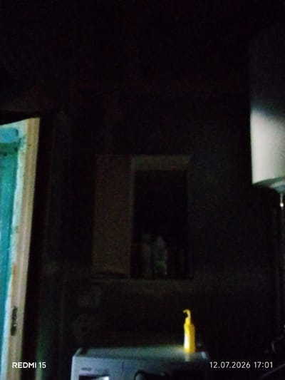 The image shows a dimly lit interior scene, possibly a room. On the left, a portion of a doorway or frame with teal and white colors is visible. In the center, there is a small, dark opening, possibly a window or alcove, within a dark wall. Below this opening, on a dark surface, sits a bright yellow pump bottle. In the lower right corner of the image, a timestamp reads "12.07.2026 17:01". In the lower left corner, text reads "REDMI 15". The overall impression is one of low light conditions. The location of Айет, Kazakhstan is not discernible from the image itself.