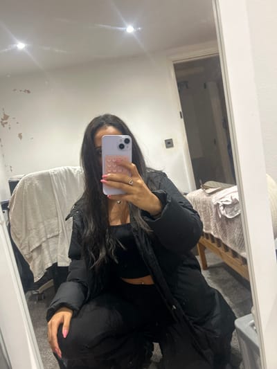 An individual is depicted taking a mirror selfie in an indoor residential setting. The specific location in Winslow, United Kingdom, cannot be determined from visual information within the image.
The subject is a female with long, dark hair, positioned in a crouch or squat. Her face is partially obscured by a light purple smartphone, held in her right hand, which features a pink circular 'pop-it' style case. Her left hand, with pink manicured nails, is visible resting on her leg. She wears a black puffer jacket over a black ribbed crop top, revealing a navel piercing, and black baggy trousers. A silver ring is visible on her left ring finger, and a silver heart-shaped pendant hangs from a necklace.
The environment includes a white wall with visible areas of peeling paint/plaster on the left side. The white ceiling has two active recessed spotlights. The floor is covered with a dark grey carpet. To the left of the subject, folded light-colored textiles, possibly towels or clothing, are visible. To the right, a wooden bed frame with light-colored bedding is partially reflected. A closed door with a dark rectangular wall fixture, possibly a light switch, is also visible in the mirror's reflection.