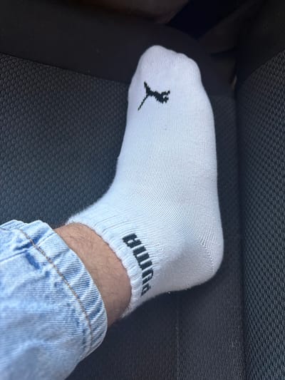 A close-up, eye-level shot focuses on a white ankle sock with a black Puma logo on the top of the foot and the word "Puma" in black lettering on the ankle band. The sock is on a person's left foot, which is partially visible above light blue denim jeans. The foot and sock are positioned on a dark, textured surface, likely a car seat, in Sculeni, Moldova. The lighting is natural, suggesting it is daytime.