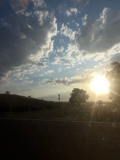 A bright sun shines through a cloudy sky over a roadside scene. To the right, a paved road stretches out beside a grassy verge with trees and a reflective sign. The sun's glare overwhelms part of the view, highlighting trees and the edge of the road. The focus is on the sky filled with scattered clouds, creating a bright, airy atmosphere.