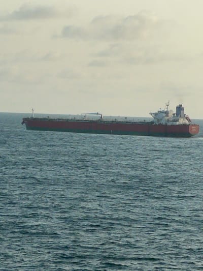 A large cargo vessel, likely an oil tanker, is depicted traversing open water under an overcast sky, observed off the coast of Akodo, Nigeria. The ship, oriented horizontally and moving from left to right, features a dark red hull with a teal or dark green boot-topping. Its stern, visible on the right, shows a white superstructure with multiple decks, radar masts, and a prominent funnel emitting a plume of dark smoke. The name "UNITY" is partially legible on the stern. Various pipes and deck structures are visible along the main deck. The sea surface exhibits moderate choppiness with visible wave patterns. The sky is a uniform, light grey, indicative of complete cloud cover. No other vessels, landmasses, or individuals are visible within the frame.