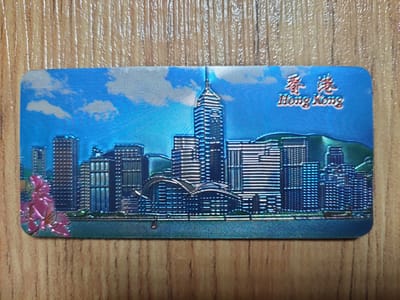 This is a close-up, top-down, eye-level shot of a holographic postcard or magnet featuring a cityscape and text. The postcard is rectangular with rounded corners and is placed on a wooden surface with a prominent wood grain pattern. The cityscape depicts several tall buildings, illuminated by what appears to be the sun, creating a bright, shimmering effect due to the holographic material. The sky above the buildings is blue with white clouds. In the upper right corner of the postcard, there are Chinese characters and the English text "Hong Kong" in a stylized font. On the left side of the postcard, near the bottom, there is a depiction of pink flowers with a metallic sheen. The postcard is positioned with its long edge running horizontally across the frame. There is no visual information within the image that relates to the city of San Pascual, Philippines.