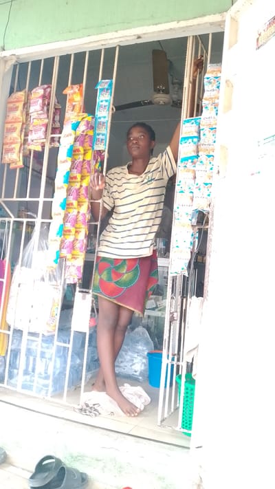A young woman stands in the doorway of a small shop in Asubi, Nigeria. The shop is protected by a metal grille. Candy and snack packets hang from the grille. The woman wears a striped shirt and patterned skirt, and she is barefoot. Inside the shop, bottled water and a ceiling fan are visible. A pair of sandals is on the ground near the shop's entrance.