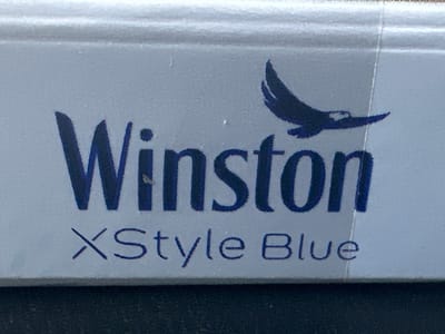 The image shows a close-up of a blue and white cigarette pack. The brand name "Winston" is printed in large, dark blue letters. Below that, in smaller, dark blue letters, is "XStyle Blue". A silhouette of a bird in flight is positioned to the right of "Winston". The background appears to be a textured surface, possibly a table or a shelf. The image quality is somewhat grainy, with some minor imperfections on the surface of the pack. The location of the image in Putilkovo, Russia, is not discernible from the visual information provided.