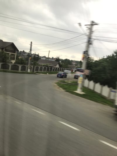 The image depicts an exterior view of a street in Cristești, Romania, captured with substantial motion blur, indicative of being taken from a moving vehicle. The sky is overcast and light grey. In the foreground, a paved road with a white lane marking slopes from the bottom left to the center-right.
On the left side of the road, a multi-story house with a brown pitched roof and a balcony is partially visible behind green trees. Extending from this area, a low, light-colored concrete wall with repeated arched cutouts runs along the road, bordering residential properties. Another house with a brown roof is visible further back, behind this wall.
In the midground, a blue car, likely an SUV or hatchback, is captured in motion, turning left off the main road onto a side path or driveway. Further to the right, a darker colored car is partially visible and blurred. A prominent, heavily blurred utility pole with numerous overhead wires creating a starburst effect stands on the right side of the road, with multiple lines extending across the upper portion of the frame. Another less distinct utility pole is present on the left side.
On the far right, a lighter-colored fence borders the road. Abundant green foliage, including trees and grass, is present throughout the scene, particularly behind structures and alongside the road. In the background, additional houses with brown roofs are visible on distant hills, suggesting a hilly or undulating landscape. No people are discernible in the image.