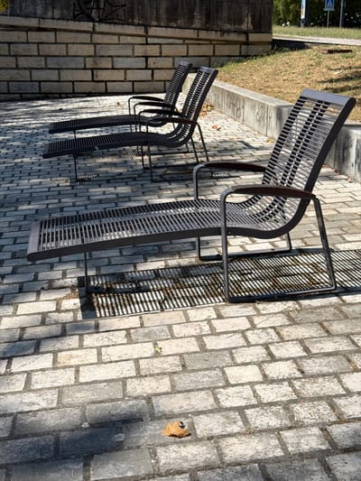 The image shows a row of empty, dark metal benches with slatted seats and backs, positioned on a paved area of light grey bricks. The benches are arranged side-by-side, and their dark frames cast striped shadows on the brickwork, indicating that the sun is shining. In the background, to the left, a grassy area with some dry leaves is visible, bordered by a concrete curb. Behind the grass, there is a hint of greenery and a road with a blue and white pedestrian crossing sign. The scene appears to be outdoors, possibly in a park or public space. There is no activity taking place, as the benches are unoccupied. The time of day is likely afternoon, given the strong shadows. The weather seems pleasant and sunny. The overall impression is one of an ordinary day in a public outdoor space, with the empty benches suggesting a moment of stillness. There is some graffiti visible on a dark stone wall to the far left of the image.