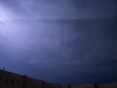 A low-angle, blurry photograph captures a dark, stormy sky with a lightning strike visible on the left side. The lower portion of the image shows the indistinct silhouette of buildings, suggesting an urban environment at night or during a severe weather event. The color palette is dominated by dark blues and purples, with the white-blue flash of lightning providing the only bright element. It is impossible to determine the specific location within Świdnica, Poland, or any details about people or their actions due to the low resolution and blurriness of the image. The scene evokes a sense of impending or ongoing storm activity over a city.