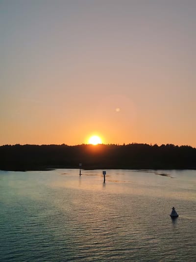 The image shows a sunset over water with a treeline in the background. Several navigational markers are visible in the water. The location is not apparent from the image.
