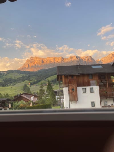 The image depicts a mountainous landscape with a village nestled in a valley. The sun is setting, casting a warm, golden light on the rugged peaks of the Dolomites. The sky is a soft blue, with scattered white clouds.
In the foreground, a modern building with wooden accents and white walls is visible. It has several windows and balconies, suggesting it might be a hotel or residential building. A small satellite dish is mounted on the roof of an adjacent structure.
Below the building, the landscape transforms into a lush green valley dotted with smaller houses and trees. The village appears to be spread out along the valley floor, with pathways and some scattered umbrellas visible in an outdoor seating area. The overall scene evokes a sense of tranquility and natural beauty, characteristic of the alpine region.
The image is oriented vertically, giving a sense of height and depth to the mountainous panorama. The lighting suggests it's late afternoon or early evening, a time often associated with serene views and the changing colors of the sky and mountains.