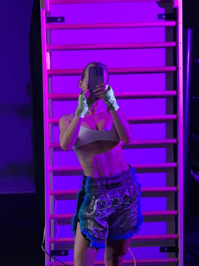 In a dimly lit indoor setting, bathed in vibrant purple lighting, a person is captured mid-action as they take a selfie. The subject, dressed in athletic attire consisting of a white sports bra and shiny, silver Muay Thai shorts with blue fringed trim, is positioned horizontally, possibly suspended or in a contorted pose against a wooden structure resembling a cage or a ladder. Their hands are wrapped in white and green tape, typical of boxing or martial arts. The individual holds a dark-colored smartphone, angled to capture their reflection in the phone's screen. The background is a wall of vertical wooden slats illuminated by the purple light, creating a dramatic and moody atmosphere. The overall impression is one of dynamic movement and a focus on athletic performance or preparation, with the selfie adding a modern, personal touch to the scene. The specific location within Kâğıthane, Turkey, is not evident from the visual cues, and the time of day or weather conditions are irrelevant given the indoor, artificially lit environment.