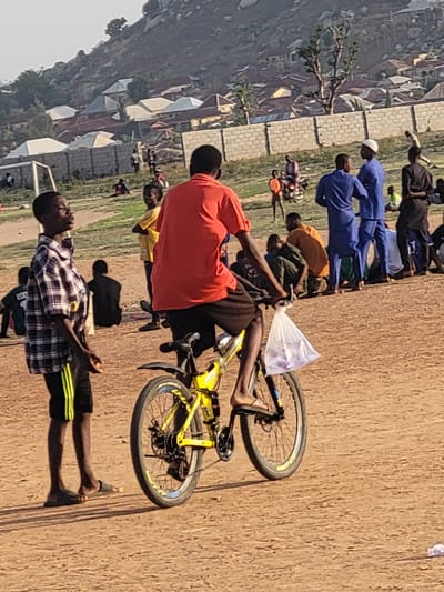 The image depicts a group of people, potentially school children, gathered in an open dirt field on the outskirts of the city of Ibo, Nigeria. A boy in a red shirt is sitting on a yellow bicycle holding a plastic bag. Other individuals are standing in a row. The background shows residential buildings and a hill. A goal post suggests the field is used for sports. Some of the children are wearing blue uniforms.