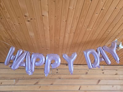 A string of rose gold foil letter balloons spelling "HAPPY DAY" is suspended from a wooden plank ceiling. The balloons are inflated and appear to be hanging from a white string. The ceiling is made of light brown wooden planks with visible grain patterns and knots. In the background, more wooden planks form a wall or the continuation of the ceiling. The lighting is neutral, casting no significant shadows. There are no people or other objects visible in the frame. The scene is static and devoid of any activity. The image is a close-up shot focusing on the balloons and the wooden ceiling. The specific location in Kapidony, Russia is not discernible from the visual information provided in the image.