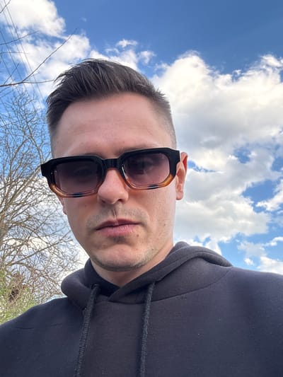 The image shows a man wearing sunglasses and a dark-colored hooded sweatshirt. He is positioned at an unusual angle, looking downwards toward the camera, with the sky and clouds visible in the background. Bare tree branches are visible at the bottom of the frame. It is unclear if the image was taken in Gorna Grashtitsa, Bulgaria. There are no definitive elements in the photograph to determine its specific location.