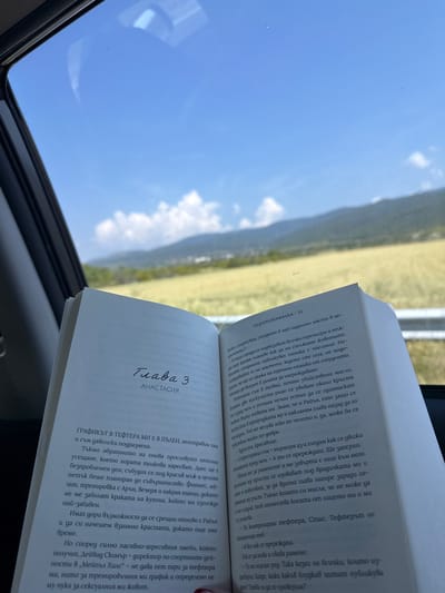 The image captures an open book in the foreground, partially obstructing a view through a vehicle window. The book displays text in Bulgarian Cyrillic. The left page is titled "Глава 3" (Chapter 3) with "АНАСТАСИЯ" (ANASTASIA) below it. The right page features the header "ЛЕДОРАЗВИВАЧКА | 35" (The Icebreaker | 35). Through the window, a bright blue sky with scattered white cumulus clouds is visible above a distant landscape. This landscape consists of green fields and rolling hills or mountains. Faint structures are discernible on the distant terrain. The car's dark window frame is visible on the left. The scene depicts a clear, sunny daytime environment, consistent with a location such as Chintulovo, Bulgaria. No individuals are visible.