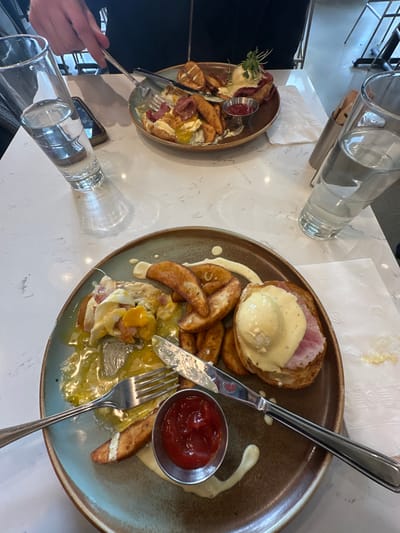 Two plates with food rest on a white table next to glasses of water. One plate displays what appears to be eggs Benedict with potato wedges and a small bowl of ketchup. A fork and knife are positioned near the eggs. The second plate mirrors the first, albeit with less of the dish visible, and features similar components arranged differently, including a garnish of greenery. A hand holding a fork is visible in the lower left corner. A cell phone is also next to the second dish.
