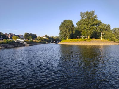 The image depicts a tranquil scene along the Rhine River in Niederkassel, Germany, on a clear, sunny day. The foreground is dominated by the dark blue, slightly rippled water of the river, reflecting the clear sky. On the left bank, a residential area is visible with several houses and trees lining the waterfront. A white ramp structure, possibly a boat dock or pier, extends into the water from the left bank. Further in the distance on the left, a lock system with several boats is present. The right bank of the river is characterized by a grassy, elevated area dotted with trees and a small gazebo or shelter. A red and white striped pole, likely a navigational marker, stands prominently on this side. The overall impression is one of a peaceful riverside landscape, likely a popular spot for recreation and boating.