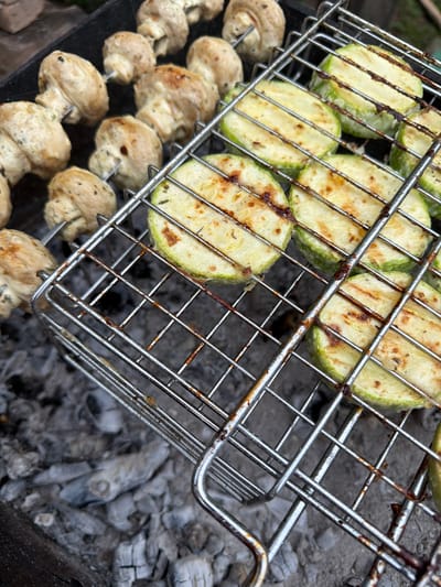 A grill basket filled with sliced zucchini and mushrooms is placed over hot coals. The zucchini slices are round and have grill marks, indicating they are being cooked. Some of the zucchini has a slightly charred appearance. Below the zucchini, there are skewers of whole mushrooms. The mushrooms are light brown and appear to be cooked as well. The grill basket is made of metal and shows signs of use. The background consists of glowing coals and ash, suggesting an outdoor cooking setting. The overall impression is a barbecue in progress. The lighting appears to be natural daylight, and there are no people visible in the image. The scene is likely taking place in a rural or backyard setting in Ильчимбетово, Russia, given the context.