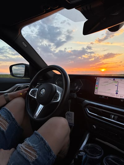 The photograph is taken from inside a car, capturing a view through the windshield toward a sunset. The dashboard, steering wheel, and center console are prominently visible, and the car's navigation screen displays a map with a route planned. The map indicates that the destination is 89 kilometers away and is near a place called Градина in Bulgaria. The estimated time of arrival (ETA) is 21:26. A person is seen wearing denim shorts, with the fabric torn near the knee. The steering wheel is held in one hand. The sky visible through the windshield shows a mix of clouds and the setting sun.