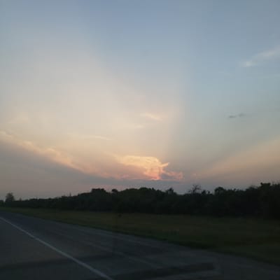 The image depicts a rural landscape scene, likely at dusk or dawn, with a road in the foreground. The sky is a gradient of soft blues and pinks, with scattered clouds. A line of trees forms a dark silhouette against the horizon. There are no people or specific structures visible, indicating an open, natural environment. The scene is devoid of any urban elements, making its association with the city of Petrovka, Russia, incongruous with the visual evidence.