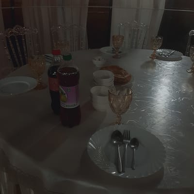 The image depicts a dimly lit dining table set for a meal. The table is covered with a white tablecloth that has a swirling, textured pattern. On the table are several place settings, each with a white plate, silverware (forks and spoons), and an amber-colored glass goblet. There are two bottles of drinks, one dark red and one dark brown, with labels that are partially visible. In the background, clear acrylic chairs are visible, some with ornate designs. There are also several small white bowls and a pastry or bread item on a plate. The overall scene suggests a formal or celebratory gathering in a dimly lit room. The location is not specified in the image.