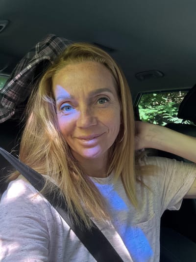 A woman with blonde hair and blue eyes is sitting in the passenger seat of a car. She is wearing a light pink t-shirt and a seatbelt. The sun is shining through the window, casting shadows and light patterns on her face and clothing. Outside the car window, green foliage is visible, indicating a natural, possibly wooded, environment. The interior of the car appears dark. The image is a selfie, taken from a front-facing perspective.
